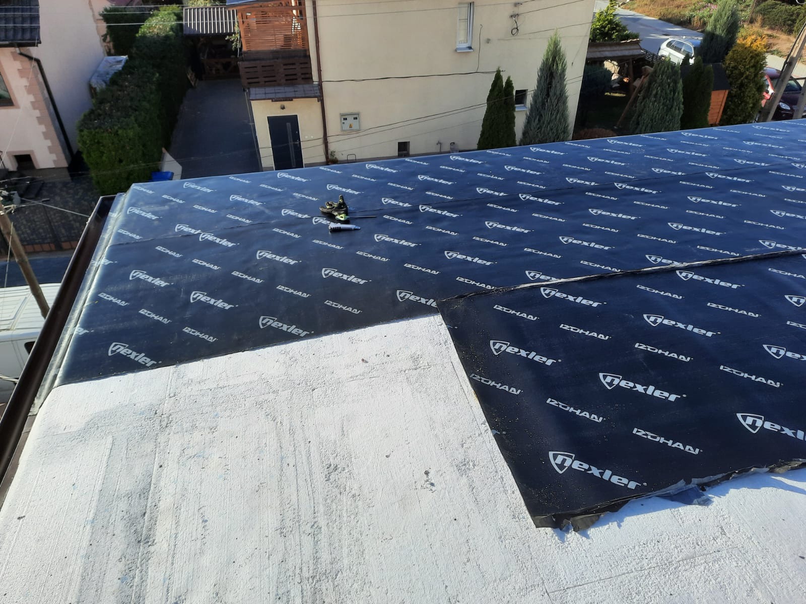 Widok z góry na fragment dachu pokrytego czarną membraną z nadrukowanym logo producenta, widoczne narzędzia dekarskie i fragment rynny, w tle budynki mieszkalne i zieleń.