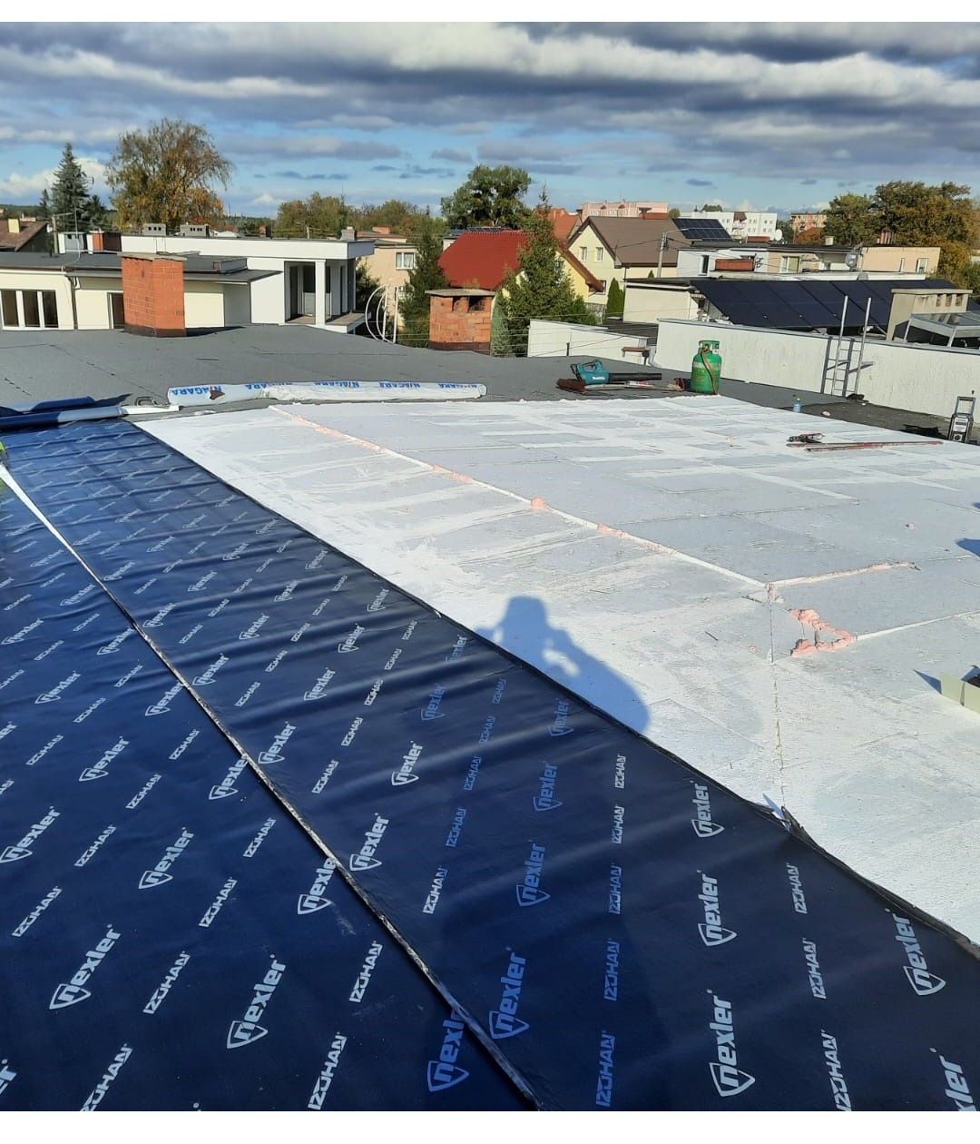 Widok z góry na płaski dach w trakcie prac: część dachu pokryta czarną membraną z logo Nexler, druga część z białym ociepleniem, w tle zabudowa jednorodzinna i błękitne niebo z chmurami.