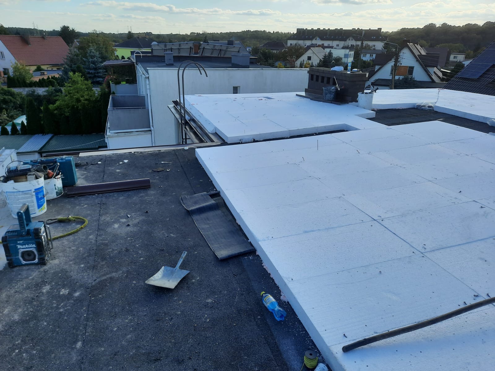 Płaski dach pokryty częściowo białym styropianem, widoczne narzędzia i materiały budowlane, w tle zabudowa miejska.