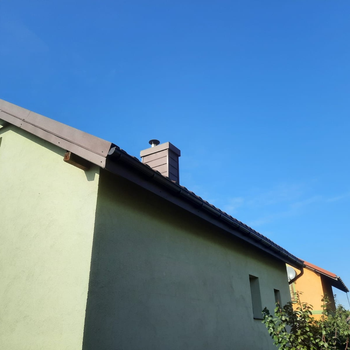 Fragment elewacji domu z widocznym kominem obłożonym panelami, rynną w kolorze ciemnego brązu i fragmentem dachu na tle błękitnego nieba.