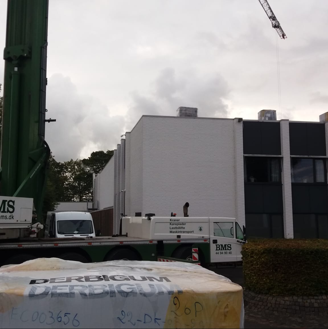 Transport materiałów budowlanych na dach za pomocą dźwigu, widoczny budynek z białej cegły i rolka papy z napisem DERBIGUM.