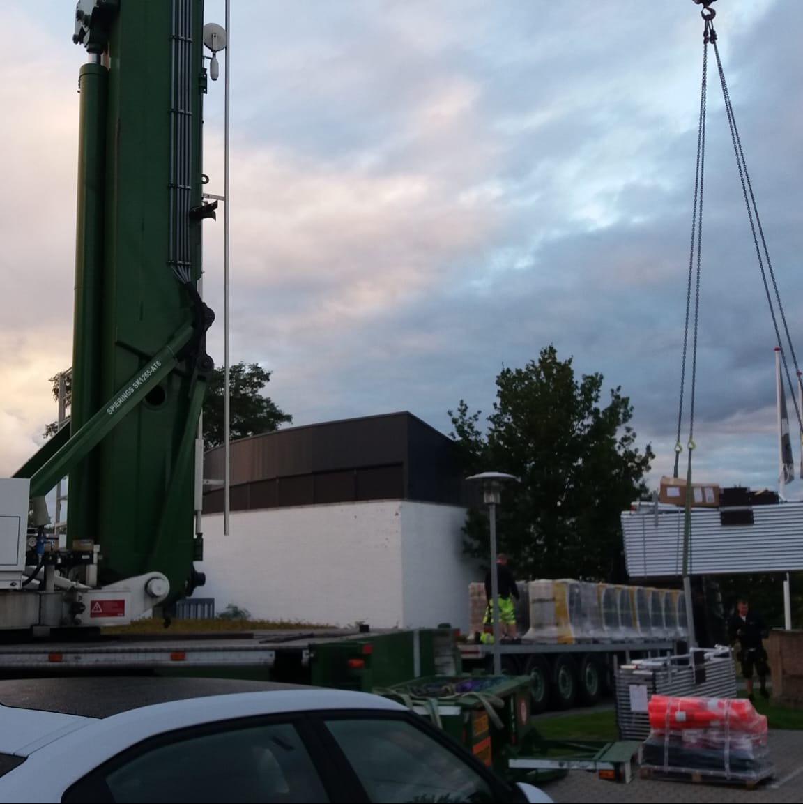 Żuraw przenosi elementy konstrukcyjne, prawdopodobnie blaszane, na tle budynku z białymi ścianami i ciemnym dachem, widoczne osoby w odblaskowych ubraniach i zaparkowany samochód.