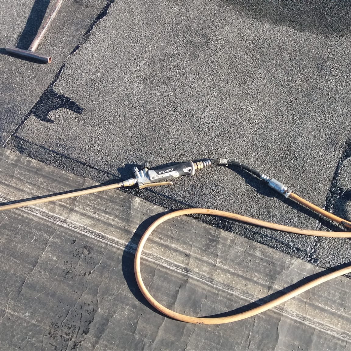 Narzędzie dekarskie Sievert z podłączonymi przewodami leży na pokryciu dachowym z papy, widoczny fragment łaty dachowej.