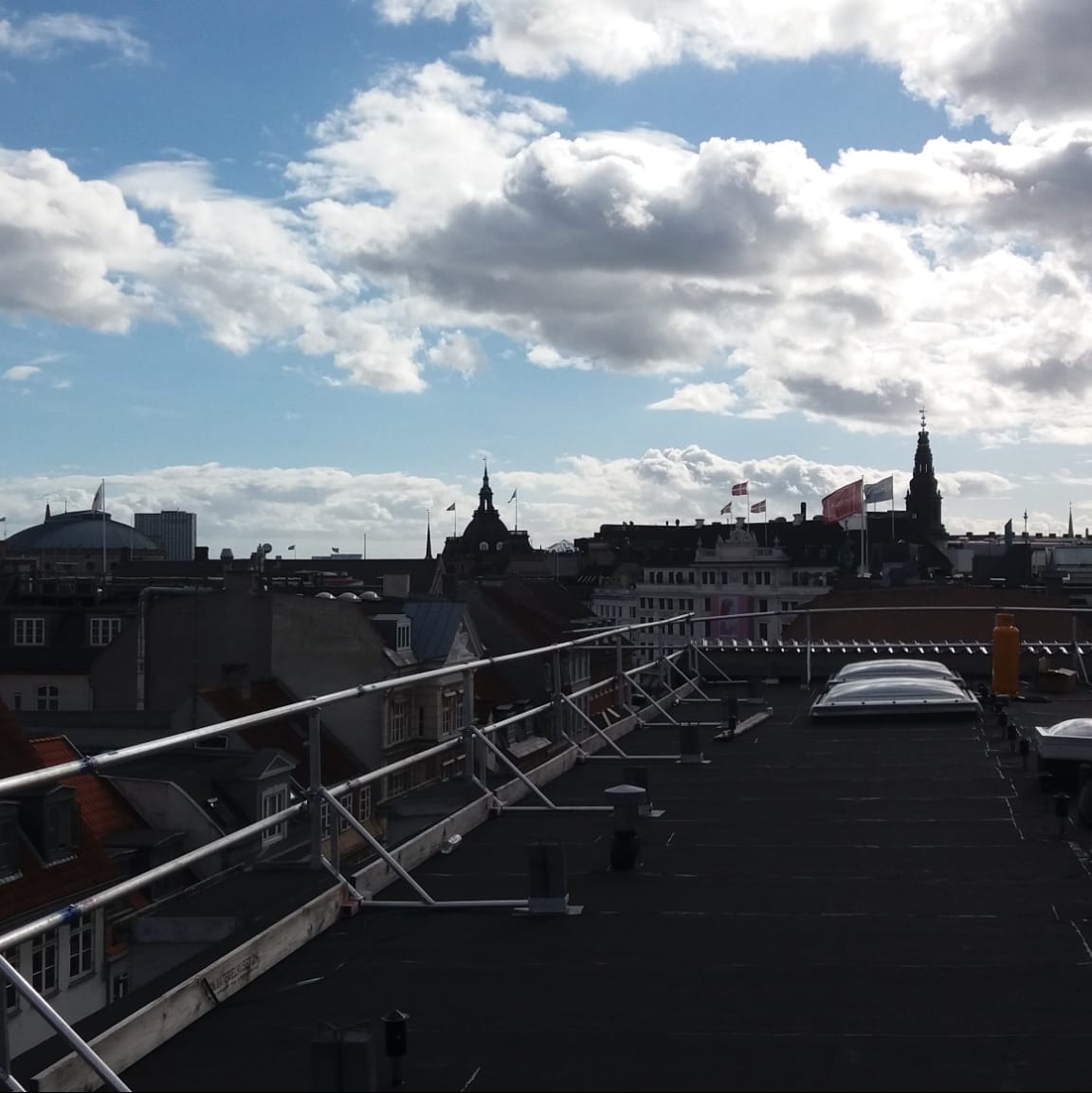 Widok z płaskiego dachu zabezpieczonego barierkami na panoramę miejską z wieżami i flagami na tle błękitnego nieba z obłokami.