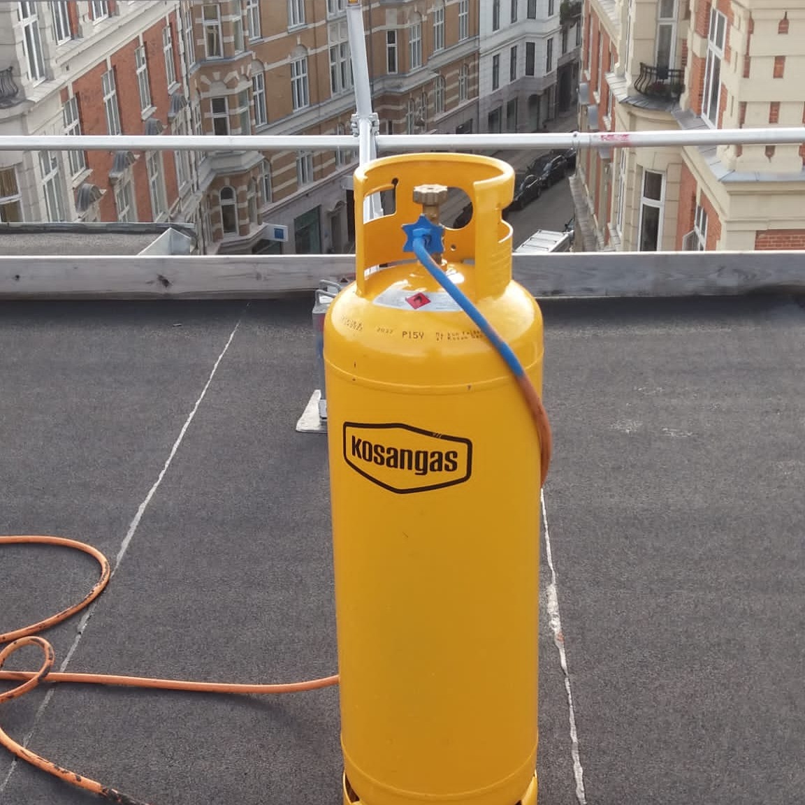 Żółta butla gazowa Kosangas z podłączonym wężem na płaskim dachu pokrytym papą, w tle budynki mieszkalne i rusztowanie.