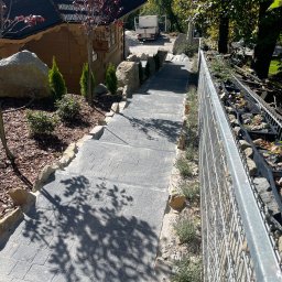 Staszel Bud - Schody z kostki brukowej prowadzące w dół, z elementami dekoracyjnymi z kamienia i roślinności, zabezpieczone metalową siatką. W tle drewniany budynek.