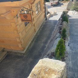 Staszel Bud - Ułożona kostka brukowa prowadząca do drewnianego domu z dekoracyjnym szyldem 'Sobiczkowa', obok kamienne obrzeża z młodymi drzewkami.