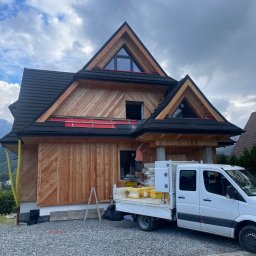 Staszel Bud - Dom w stylu góralskim w trakcie wykańczania elewacji drewnianej. Widoczny pracownik na wysokości, biały samochód dostawczy i materiały budowlane.