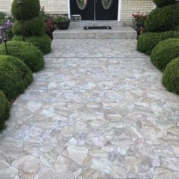 Staszel Bud - Kamienny chodnik prowadzący do drzwi wejściowych, otoczony ozdobnymi krzewami. Jasna elewacja domu w tle. Widok z góry na wzór ułożenia kamieni.