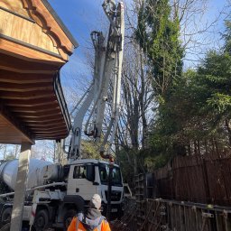 Staszel Bud - Betoniarka pompuje beton do szalunków fundamentów w Kościelisku. Pracownik w odblaskowej kamizelce nadzoruje proces. Widoczne drzewa i fragment drewnianej konstrukcji.