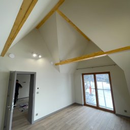 Staszel Bud - Jasne poddasze po remoncie: drewniane belki, okno z widokiem na zaśnieżony stok, panele podłogowe. Widoczny fragment osoby w drzwiach.