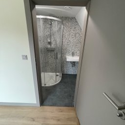 Staszel Bud - Nowoczesna łazienka z kabiną prysznicową narożną i umywalką, widoczna przez otwarte drzwi z szarą ościeżnicą. Podłoga wyłożona panelami imitującymi drewno.