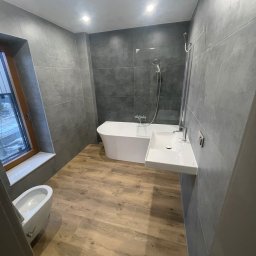 Staszel Bud - Nowoczesna łazienka z wolnostojącą wanną, wiszącą umywalką i bidetem. Szare płytki na ścianach kontrastują z drewnopodobną podłogą. Okno z widokiem na zewnątrz.