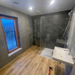 Staszel Bud - Nowoczesna łazienka z kabiną prysznicową, umywalką i drewnopodobną podłogą. Widoczne narzędzia do silikonowania. Za oknem zimowy krajobraz.