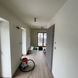 Staszel Bud - Jasne wnętrze po malowaniu ścian, widoczny odkurzacz przemysłowy, podłoga drewniana, szare drzwi i okno w tle. Minimalistyczne oświetlenie sufitowe.