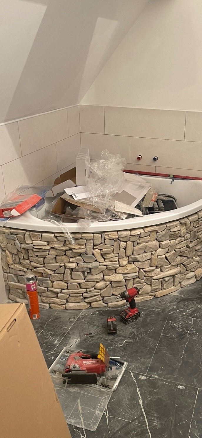 Wykończona wanna narożna z obudową z kamienia naturalnego w łazience z ciemną, marmurkową podłogą, w trakcie prac wykończeniowych. Widoczne narzędzia.