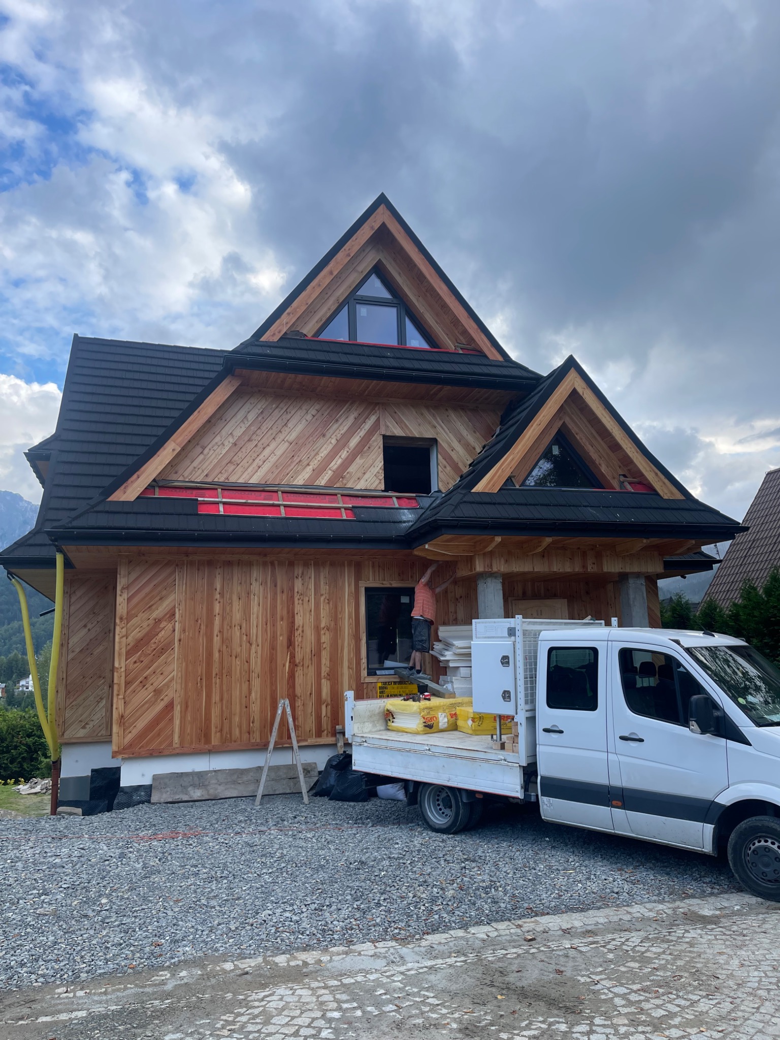 Dom w stylu góralskim w trakcie wykańczania elewacji drewnianej. Widoczny pracownik na wysokości, biały samochód dostawczy i materiały budowlane.