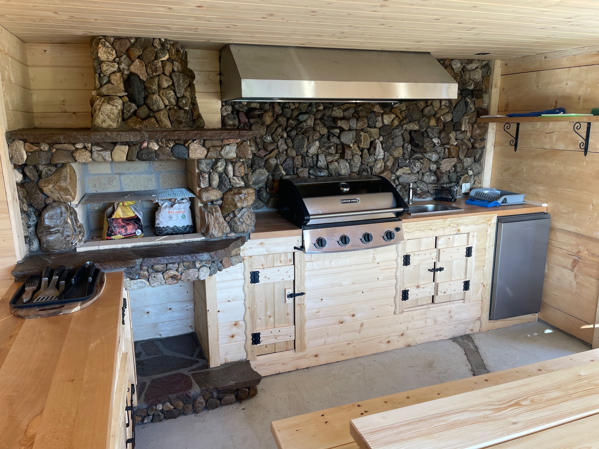 Drewniana kuchnia letnia z grillem, kamiennym kominkiem i blatem. Widoczne akcesoria do grillowania i lodówka. Całość w rustykalnym stylu, z elementami z naturalnego kamienia.