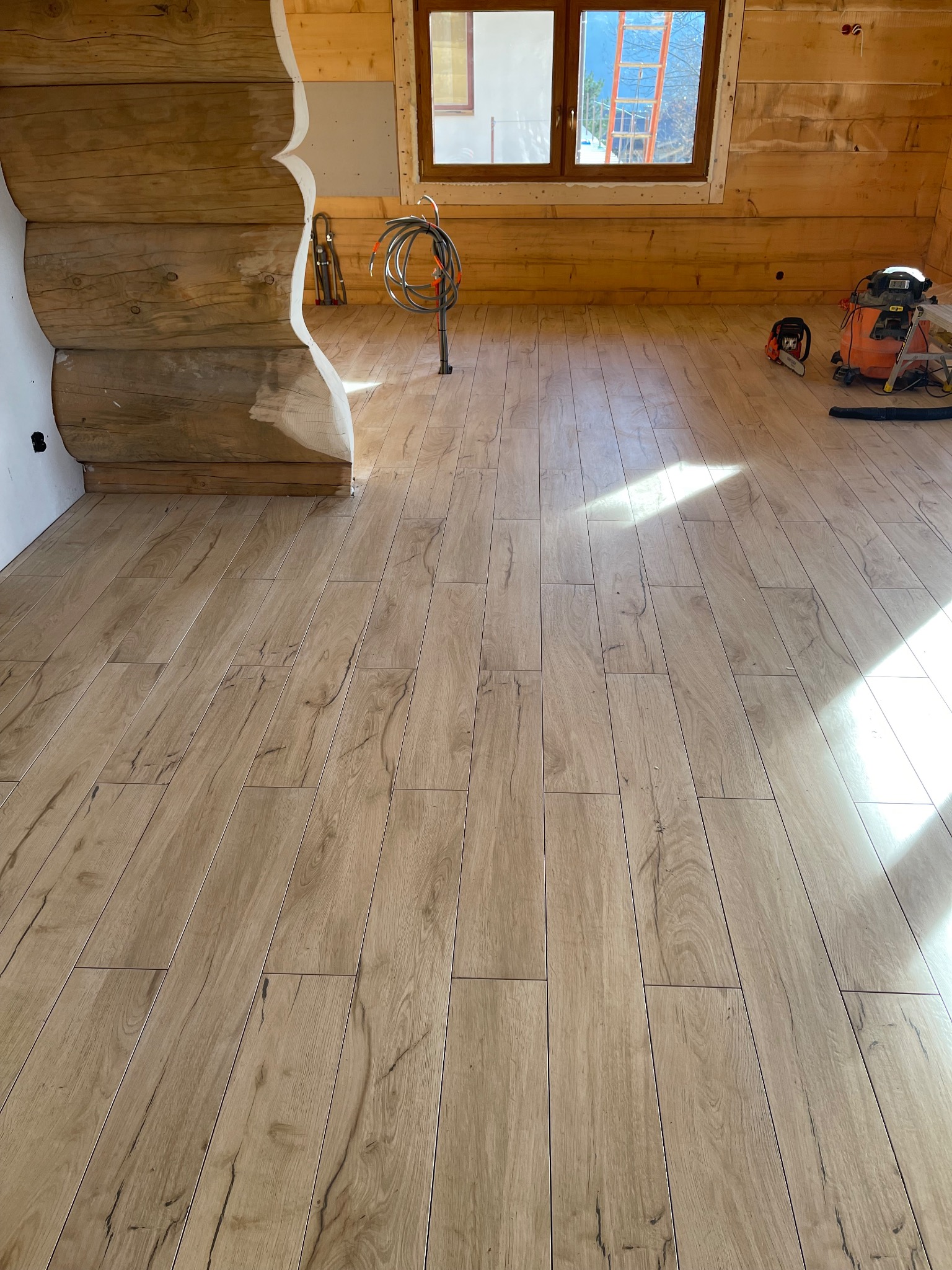 Ułożona podłoga z paneli imitujących drewno w surowym wnętrzu domu z bali. Widoczne narzędzia i kable, okno z widokiem na zewnątrz. Jasne światło dzienne.