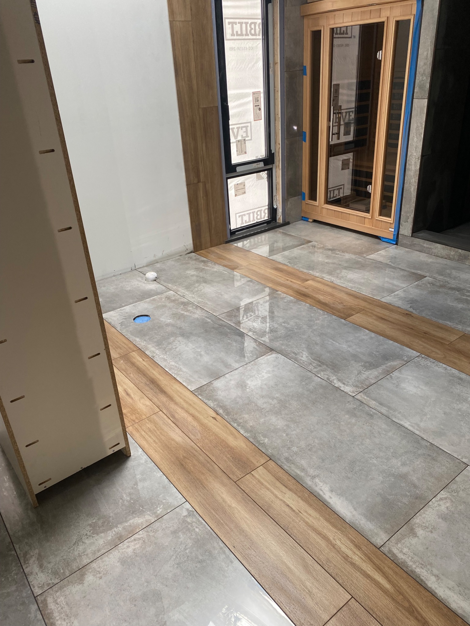 Ułożona szara płytka podłogowa z drewnopodobnymi wstawkami, fragment sauny i mebla w tle. Widoczny otwór w podłodze. Wykończenie wnętrza w trakcie realizacji.