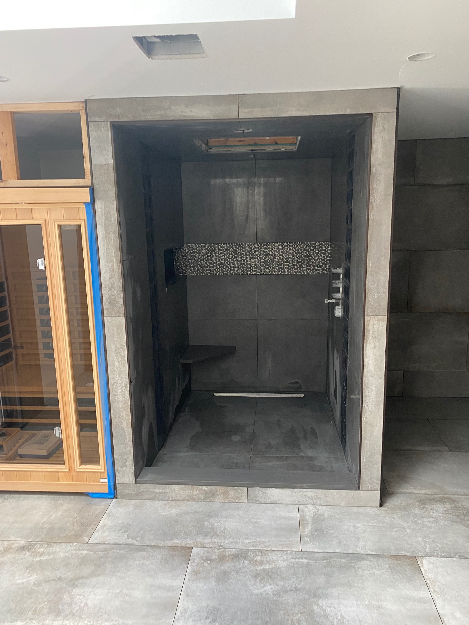 Nowoczesna kabina prysznicowa z szarymi płytkami i dekoracyjnym pasem mozaiki, obok sauna z drewnianym wykończeniem, widoczne niedokończone prace wykończeniowe.