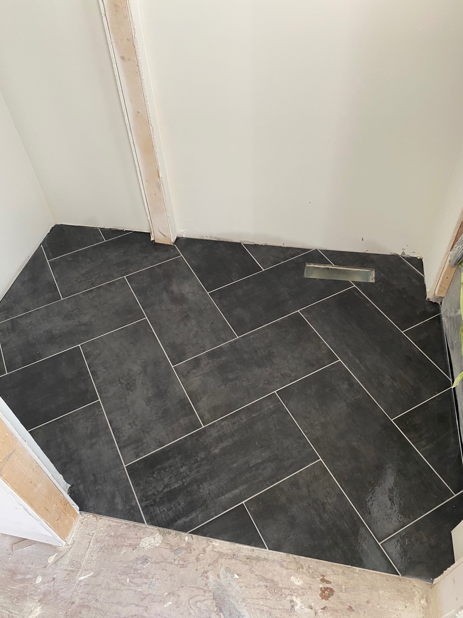 Ukończona podłoga z ciemnych, matowych płytek ułożonych w jodełkę, z jasną fugą. Widoczny fragment kratki wentylacyjnej i surowy stan wykończenia przy progu.