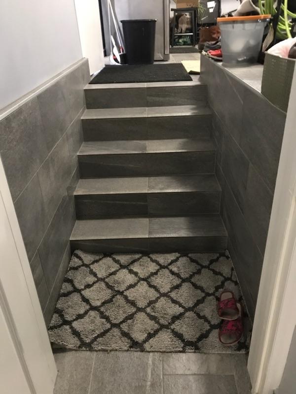 Wykończone schody do piwnicy z szarymi płytkami, dywanikiem i kapciami. Widok z góry na dół, neutralne oświetlenie. Estetyczne i funkcjonalne rozwiązanie.