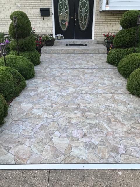 Kamienny chodnik prowadzący do drzwi wejściowych, otoczony ozdobnymi krzewami. Jasna elewacja domu w tle. Widok z góry na wzór ułożenia kamieni.