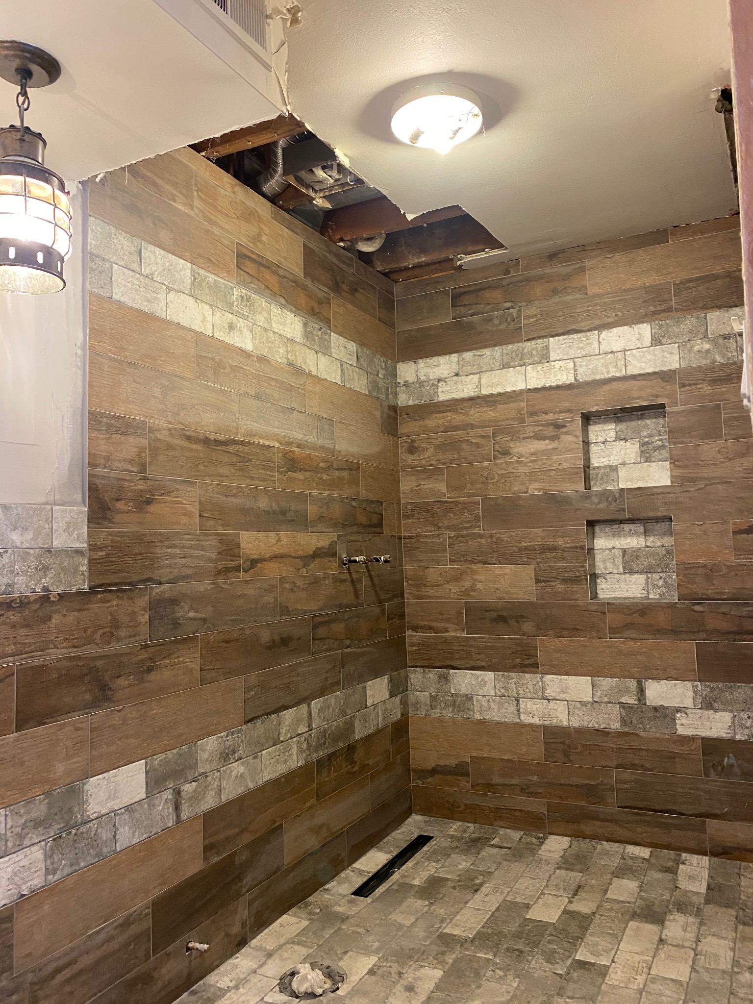 Wykończona kabina prysznicowa z drewnopodobnymi płytkami i wnękami. Widoczne niedokończone prace na suficie i instalacja oświetleniowa w stylu industrialnym.