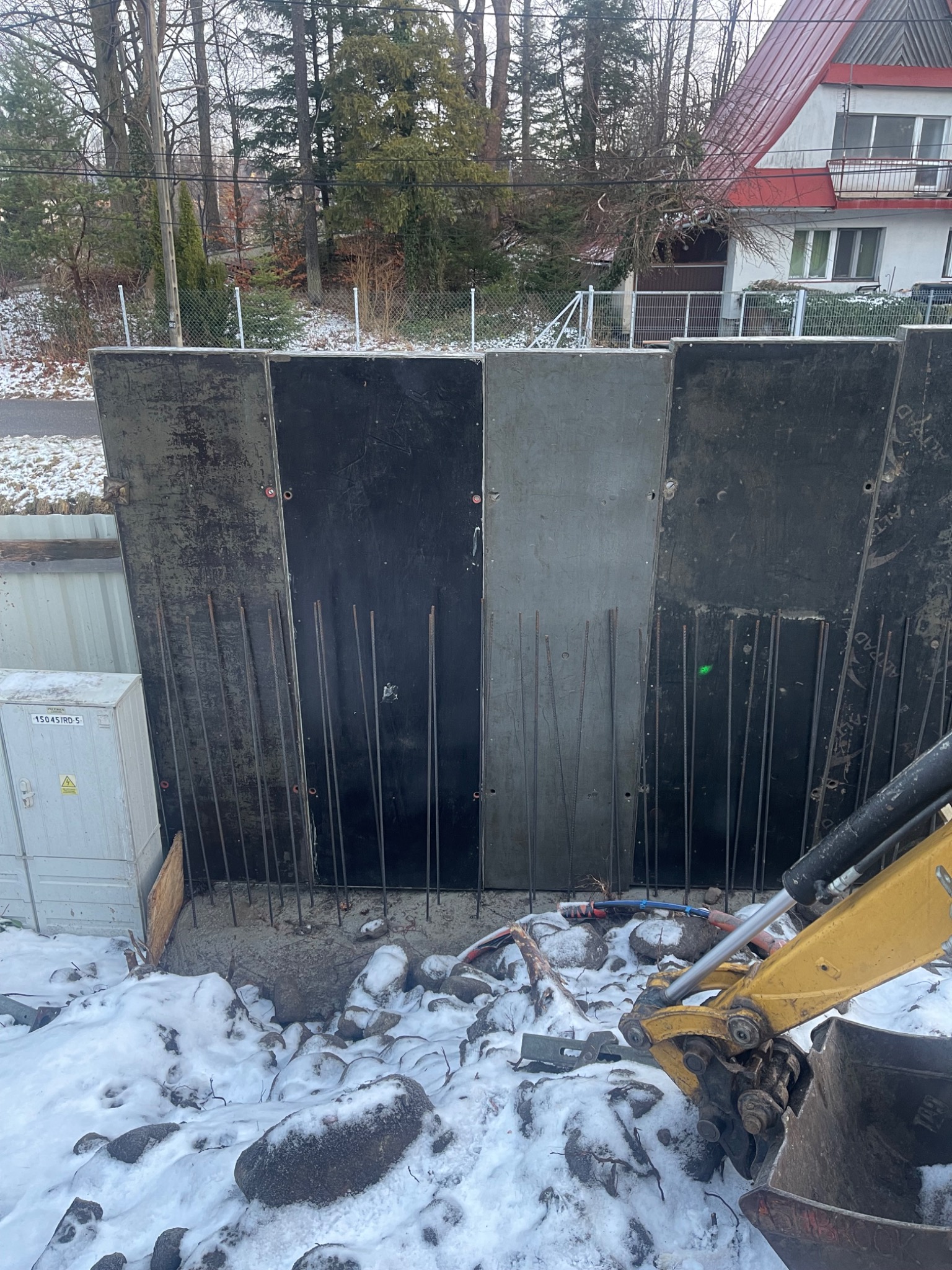 Szczegółowy widok szalunku fundamentów z wystającymi prętami zbrojeniowymi, częściowo zasypanego śniegiem, z fragmentem koparki widocznym w prawym dolnym rogu.