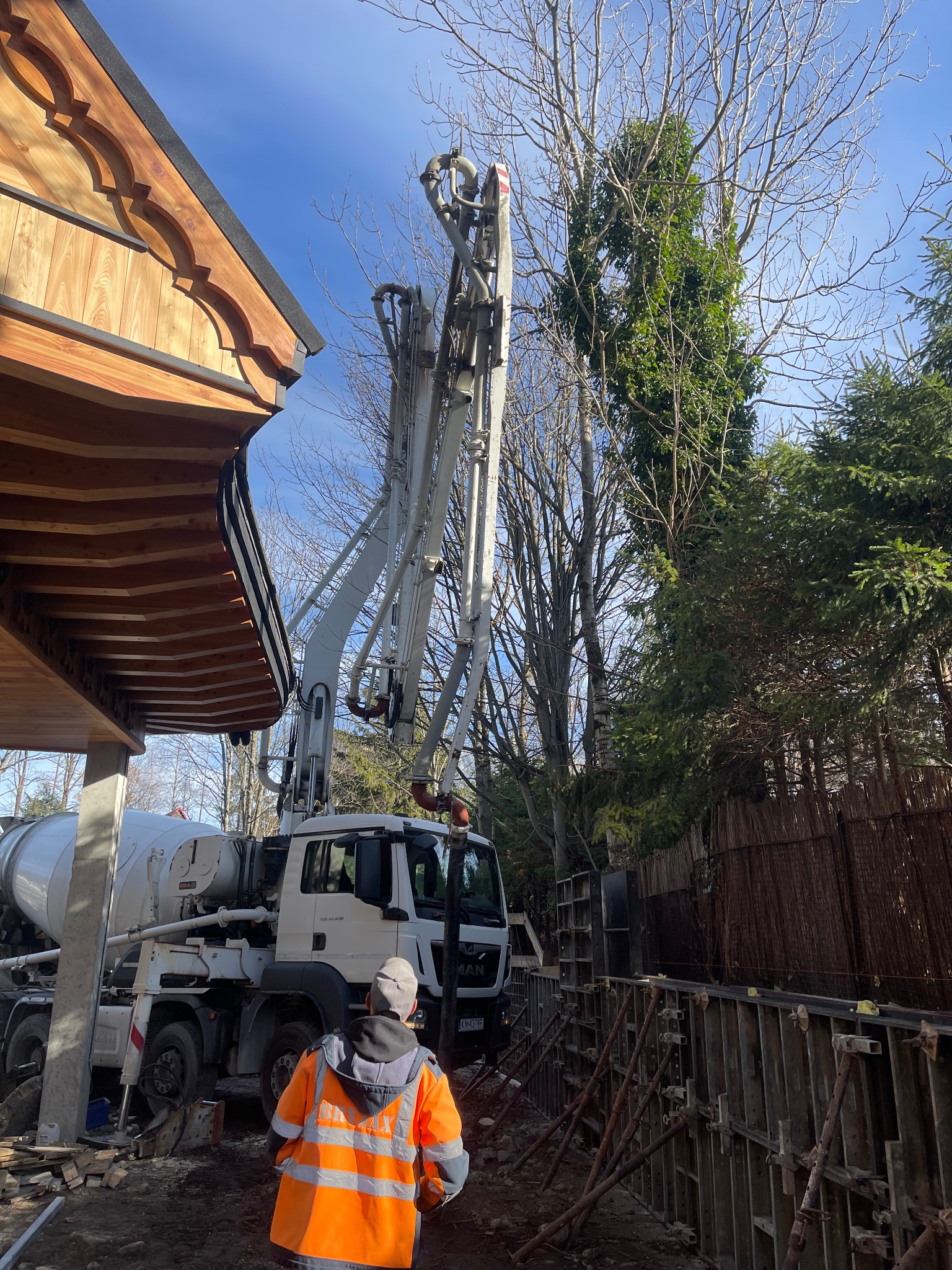 Betoniarka pompuje beton do szalunków fundamentów w Kościelisku. Pracownik w odblaskowej kamizelce nadzoruje proces. Widoczne drzewa i fragment drewnianej konstrukcji.