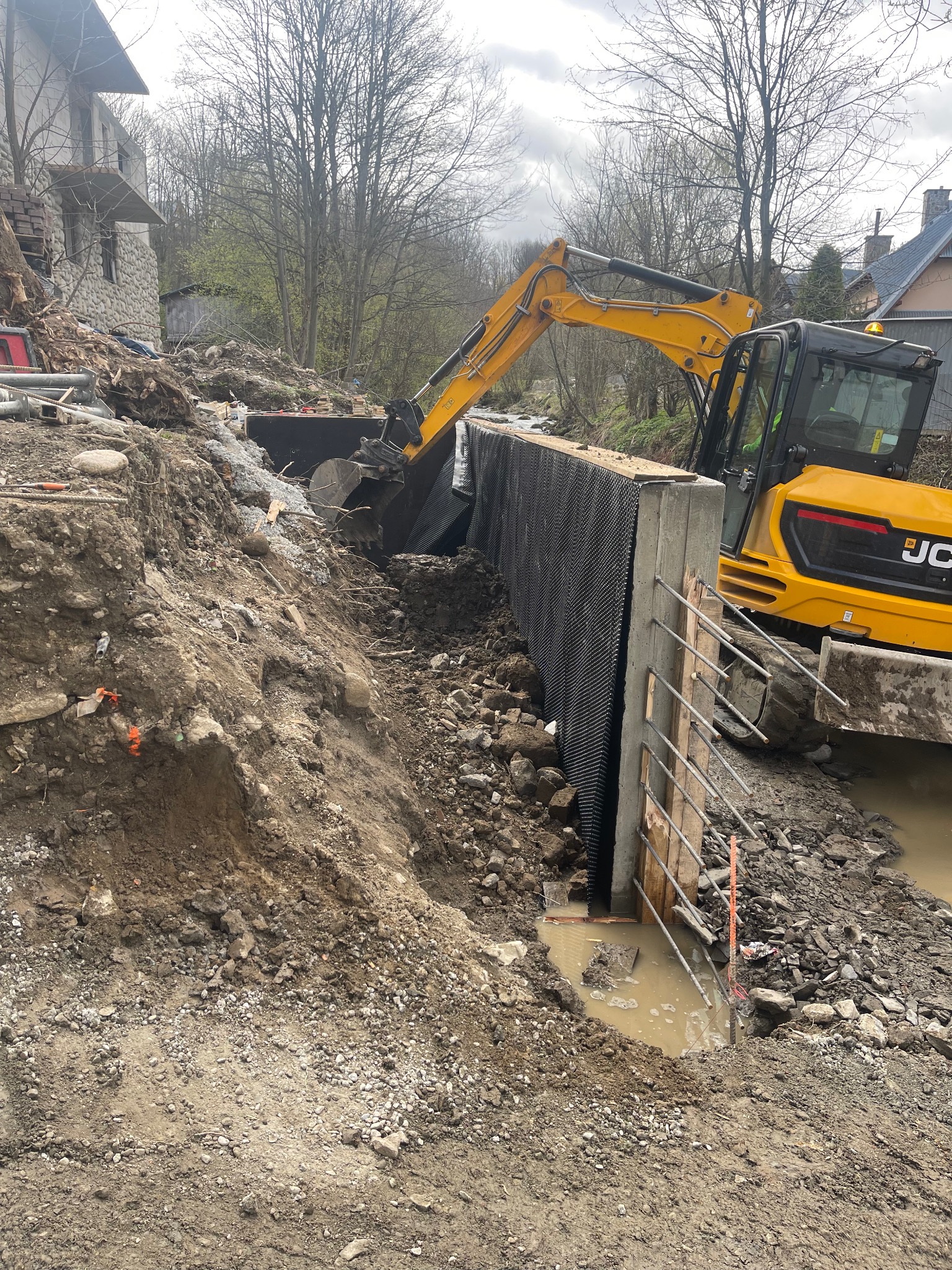 Żółta koparka JCB przy budowie betonowego muru oporowego z drenażem w wykopie, zabezpieczającego brzeg cieku wodnego. Widoczne zbrojenie i drewniane szalunki.