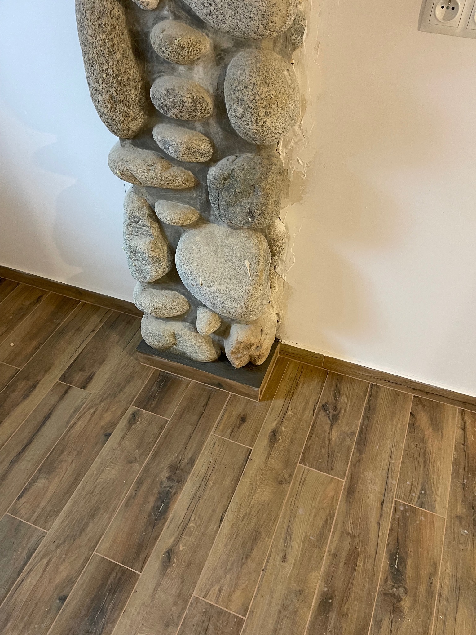 Wykończenie ściany kamieniami rzecznymi wewnątrz pomieszczenia, z widoczną podłogą z płytek imitujących drewno i fragmentem białej ściany z gniazdkami.