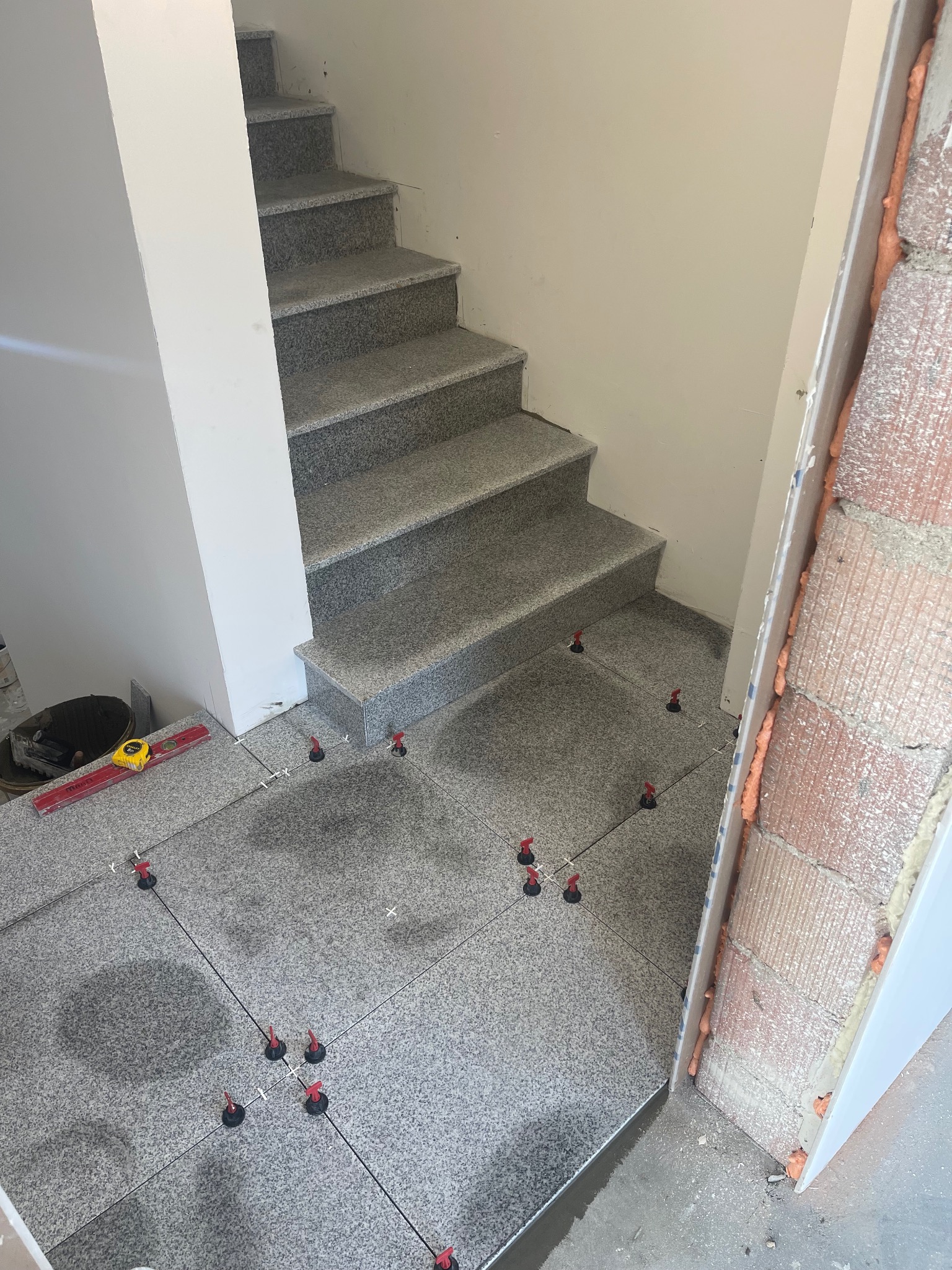 Układanie szarych, granitowych płytek na podłodze i schodach wewnątrz budynku. Widoczne narzędzia glazurnicze i system poziomowania płytek.