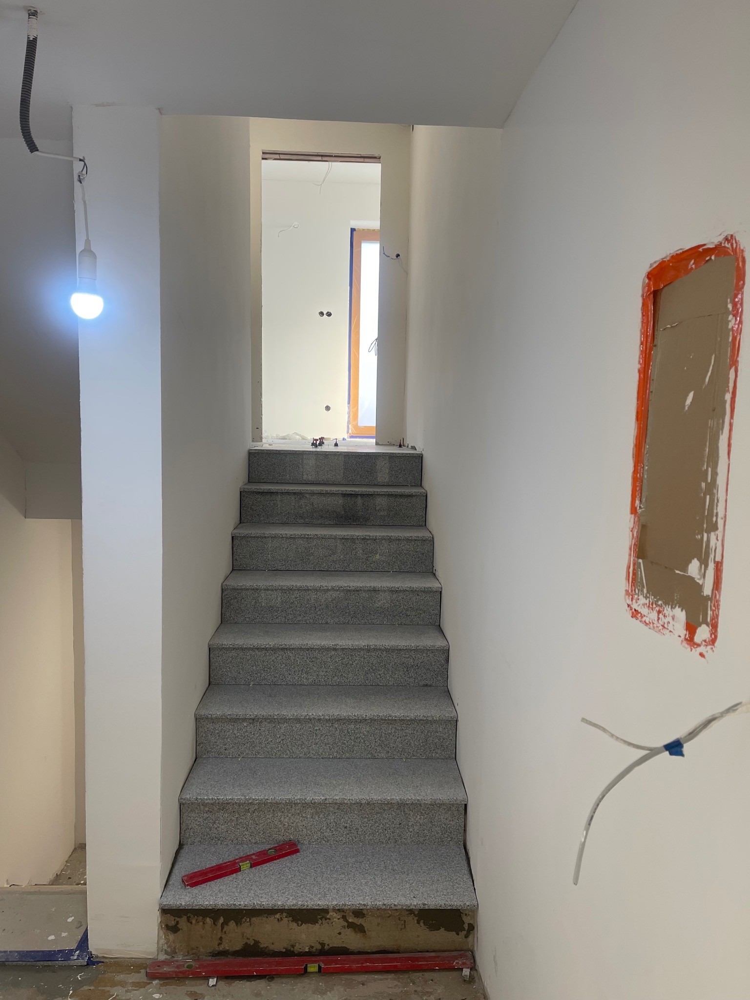 Wnętrze w trakcie wykończenia: szare schody prowadzą w górę, widoczne przygotowane podłoże, poziomica na schodach, instalacja elektryczna w ścianie i otwór po gniazdku.