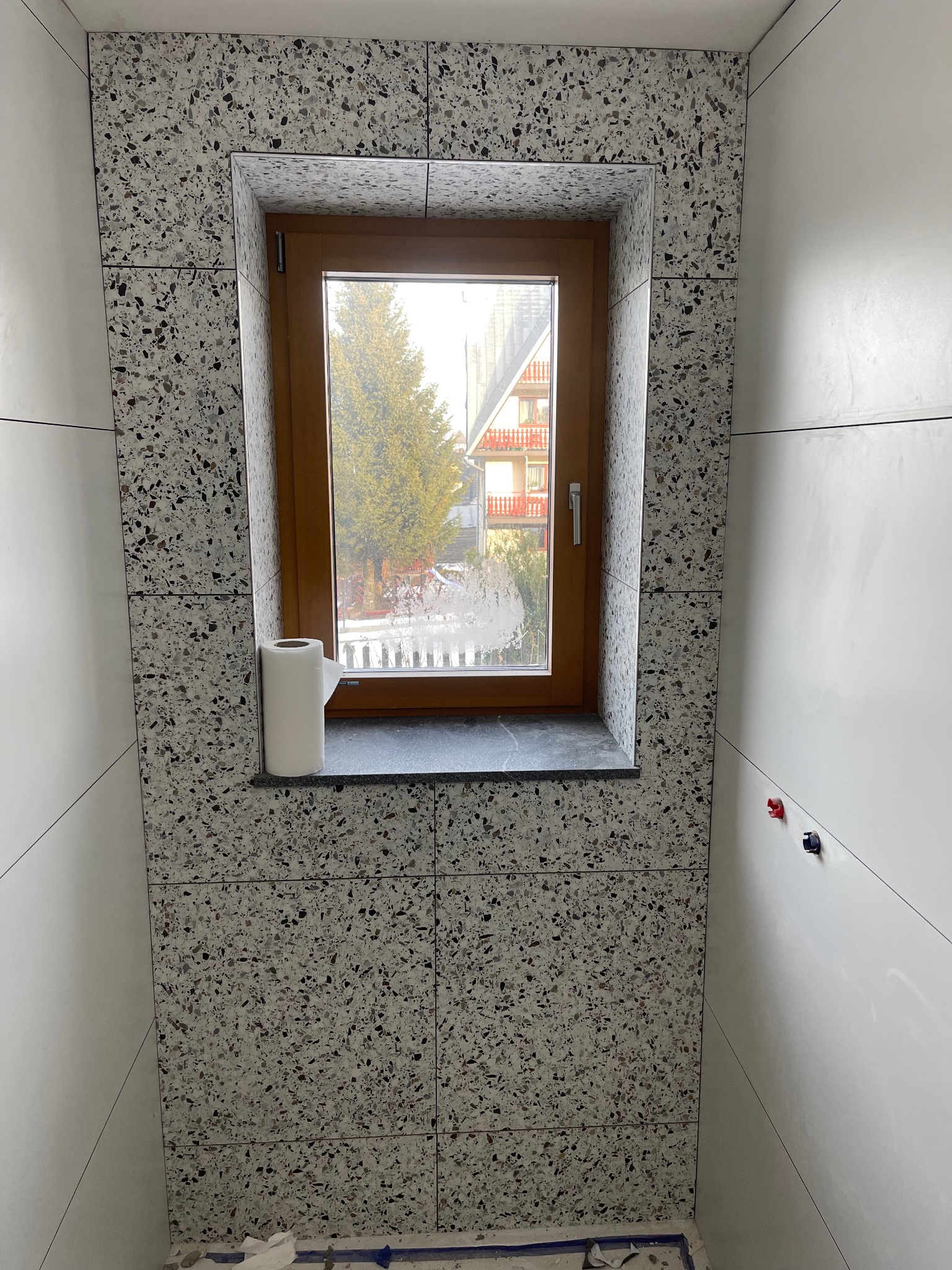 Wykończona ściana w łazience z oknem, płytki imitujące lastryko. Widoczne białe, gładkie panele ścienne i drewniana rama okienna. Na parapecie rolka papieru toaletowego.