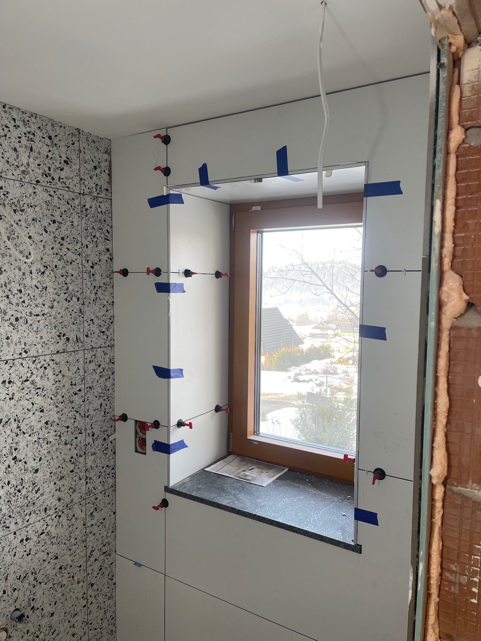 Układanie płytek w Kościelisku, widoczne narzędzia i poziomice. Ściana z oknem w trakcie wykańczania. Fragment ściany z płytkami o wzorze lastryko.