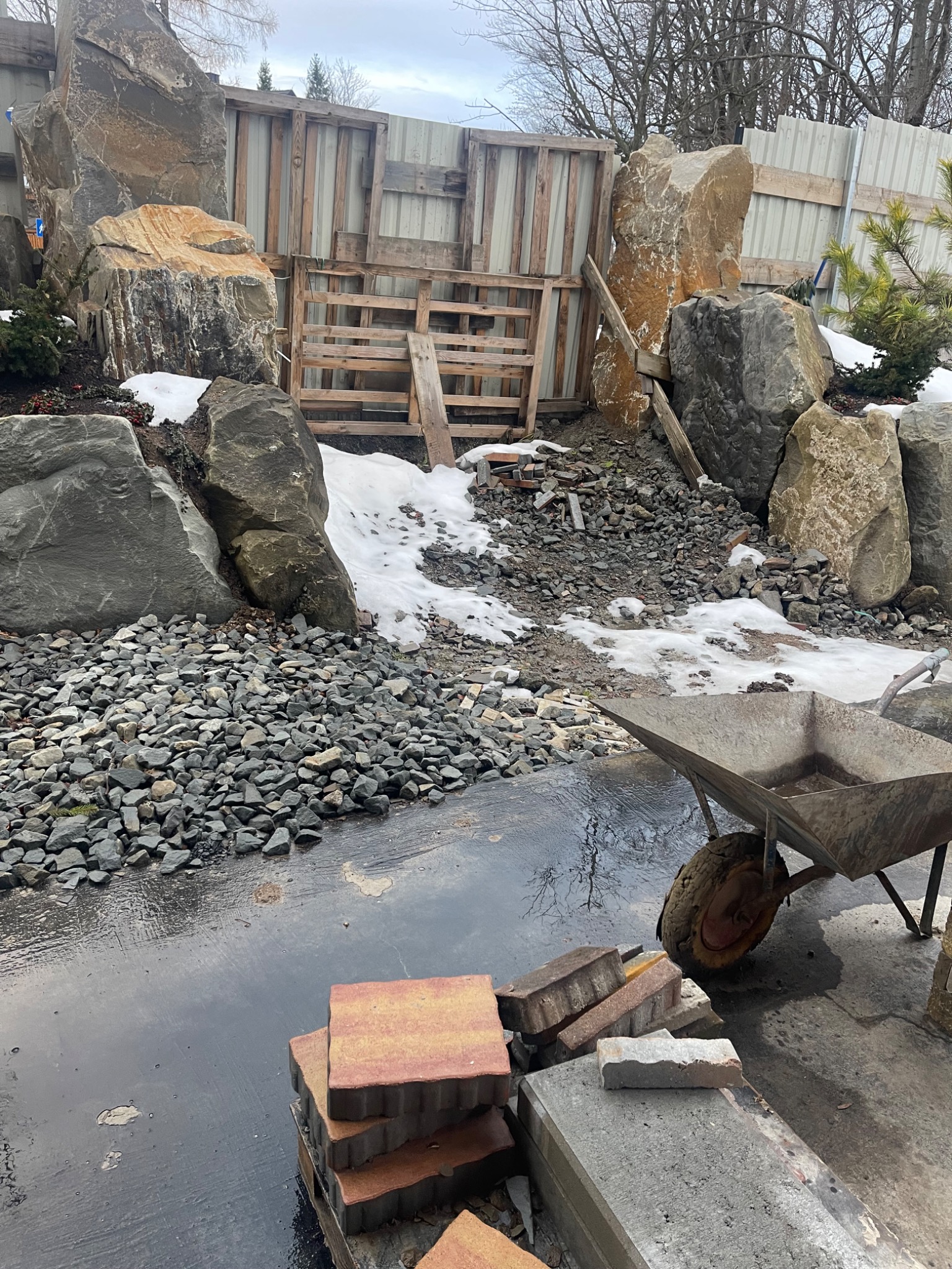Teren w trakcie prac ziemnych z kamieniami, kostką brukową, taczką i drewnianymi elementami szalunku. Widoczne ślady po deszczu i resztki śniegu.