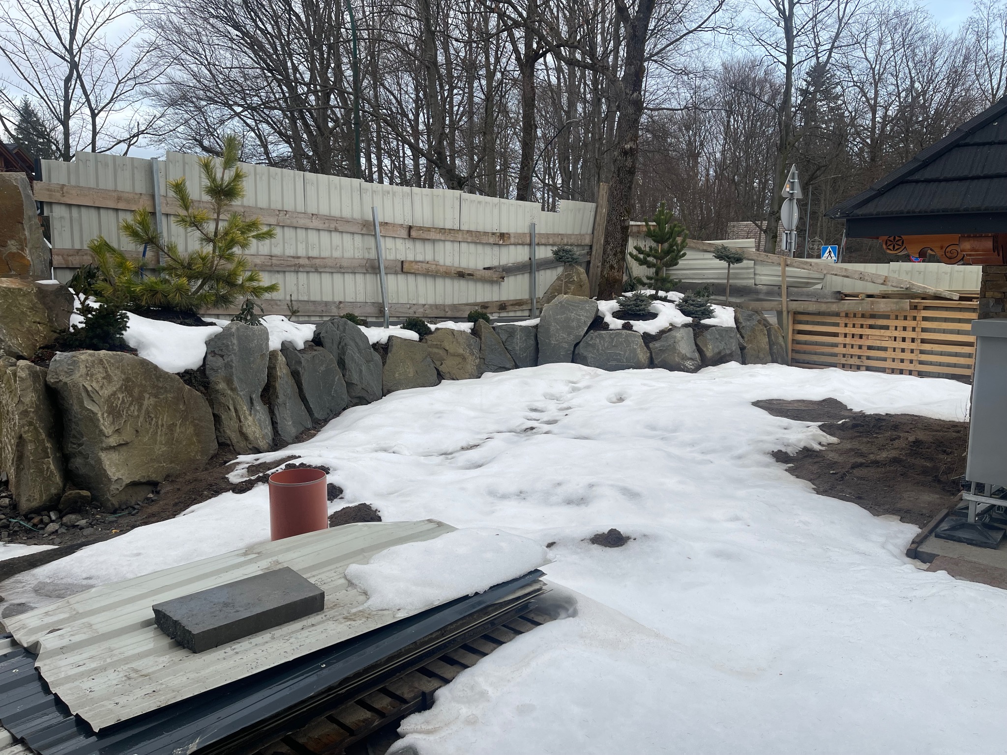 Zimowy krajobraz z kamiennym murkiem i młodymi drzewkami iglastymi, częściowo pokryty śniegiem. W tle widoczne ogrodzenie z blachy falistej i fragment drewnianej konstrukcji.