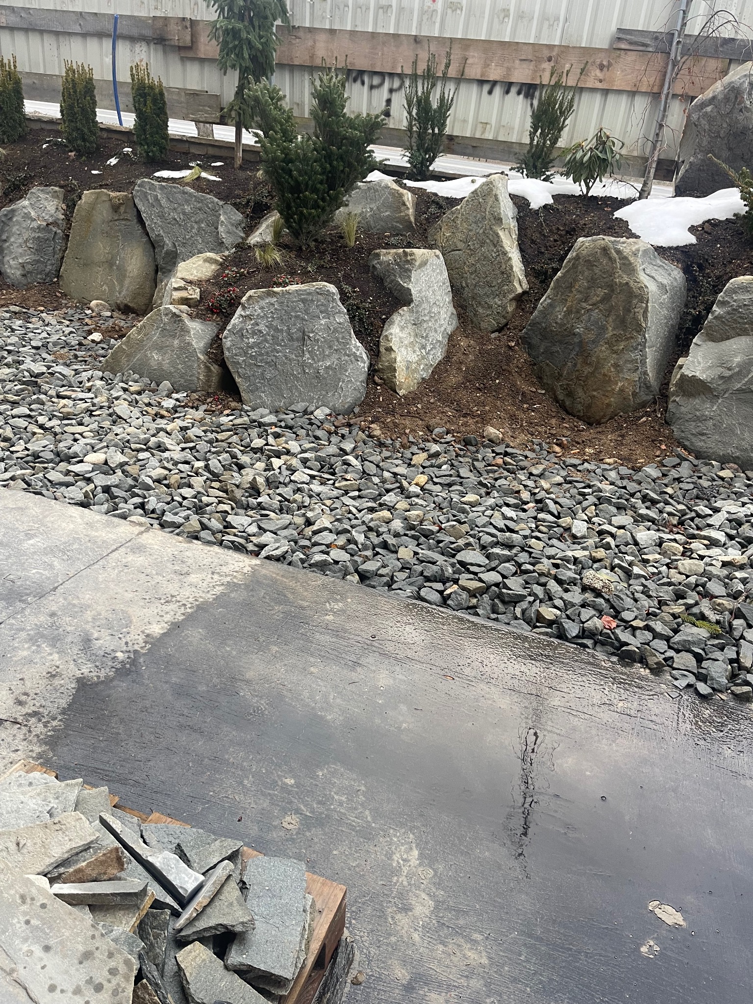 Aranżacja ogrodu z dużymi kamieniami i żwirem, świeżo po deszczu. Na pierwszym planie paleta z płytami kamiennymi. W tle roślinność i ogrodzenie.