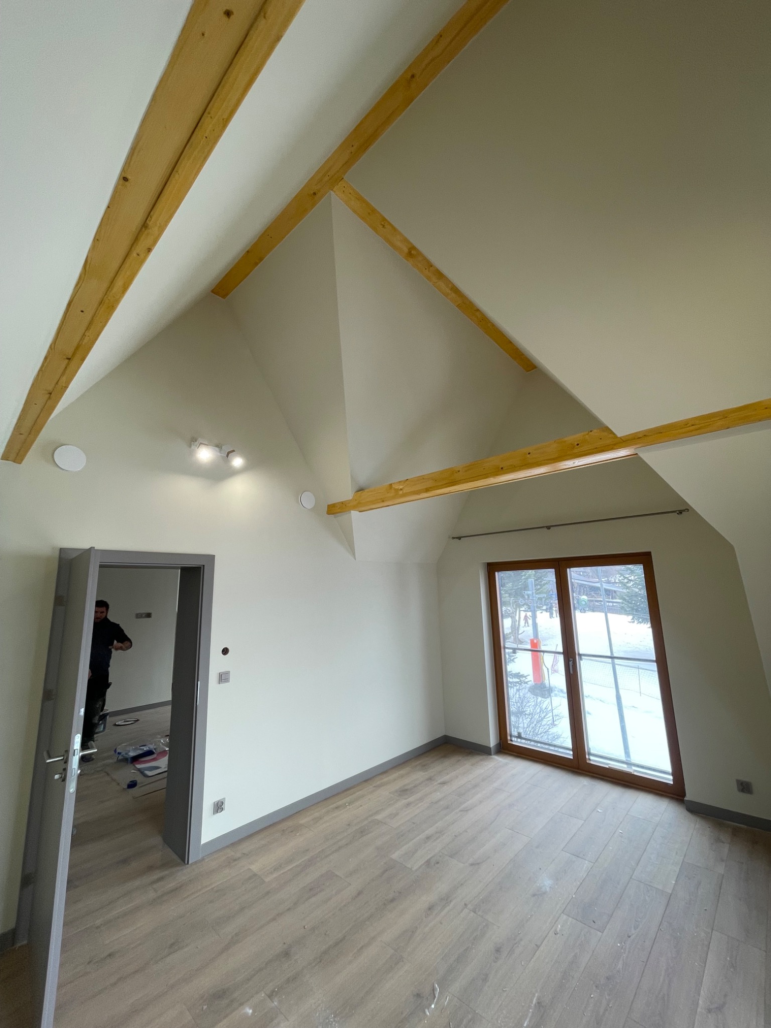 Jasne poddasze po remoncie: drewniane belki, okno z widokiem na zaśnieżony stok, panele podłogowe. Widoczny fragment osoby w drzwiach.