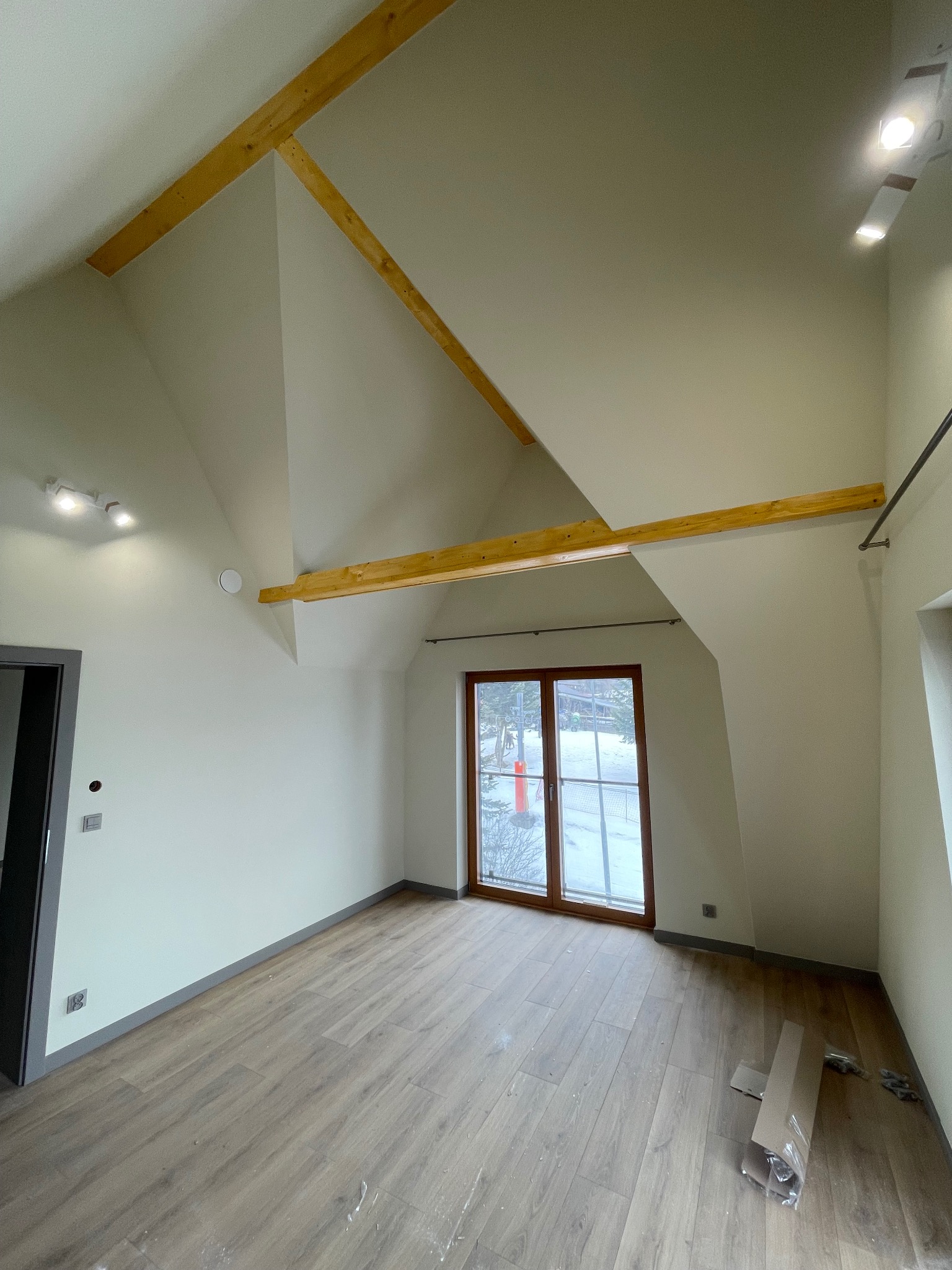 Jasne poddasze z drewnianymi belkami stropowymi i panelami podłogowymi. Okno z widokiem na zimowy krajobraz. Świeżo wykończone wnętrze z nowoczesnym oświetleniem.