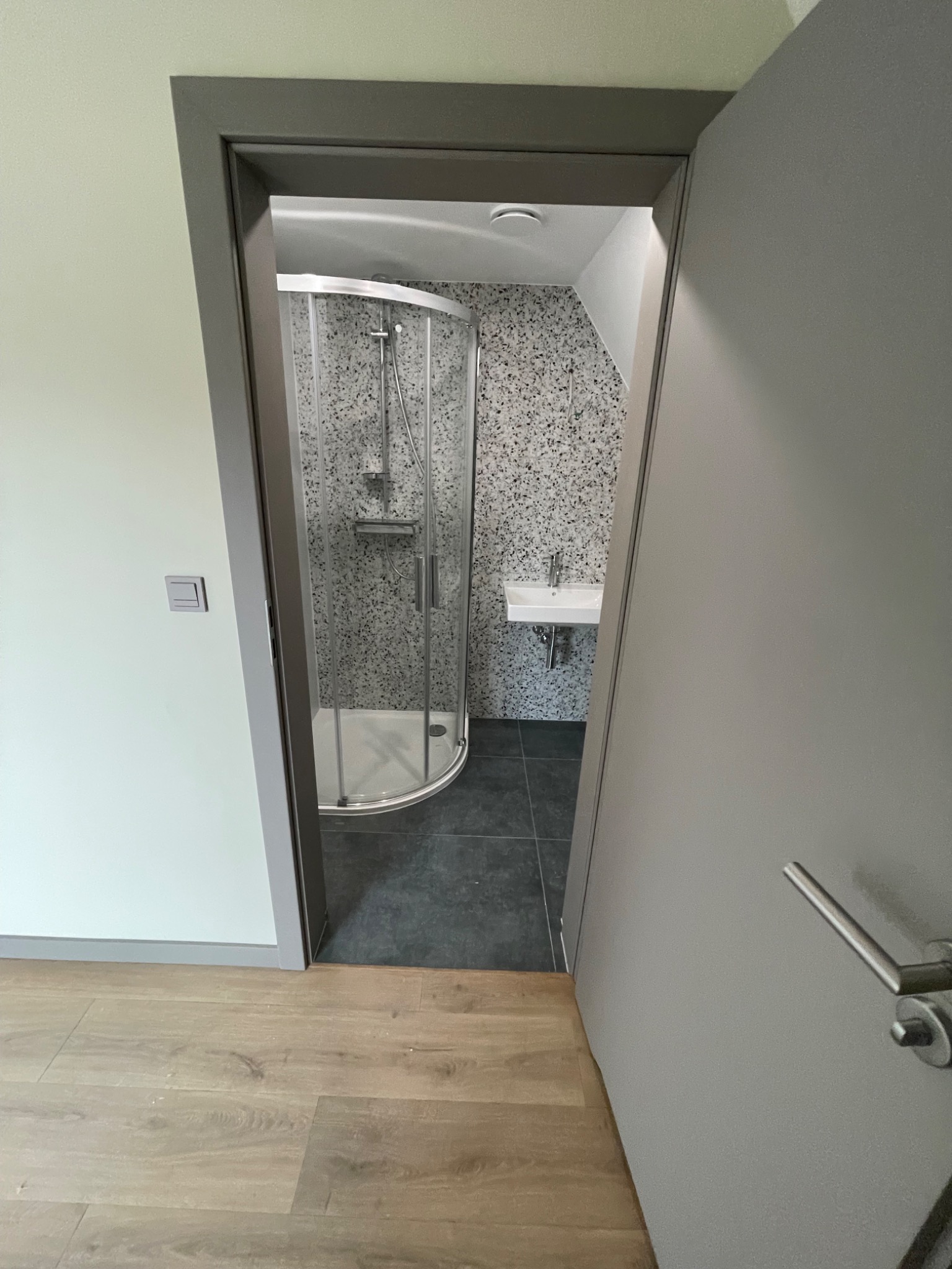 Nowoczesna łazienka z kabiną prysznicową narożną i umywalką, widoczna przez otwarte drzwi z szarą ościeżnicą. Podłoga wyłożona panelami imitującymi drewno.