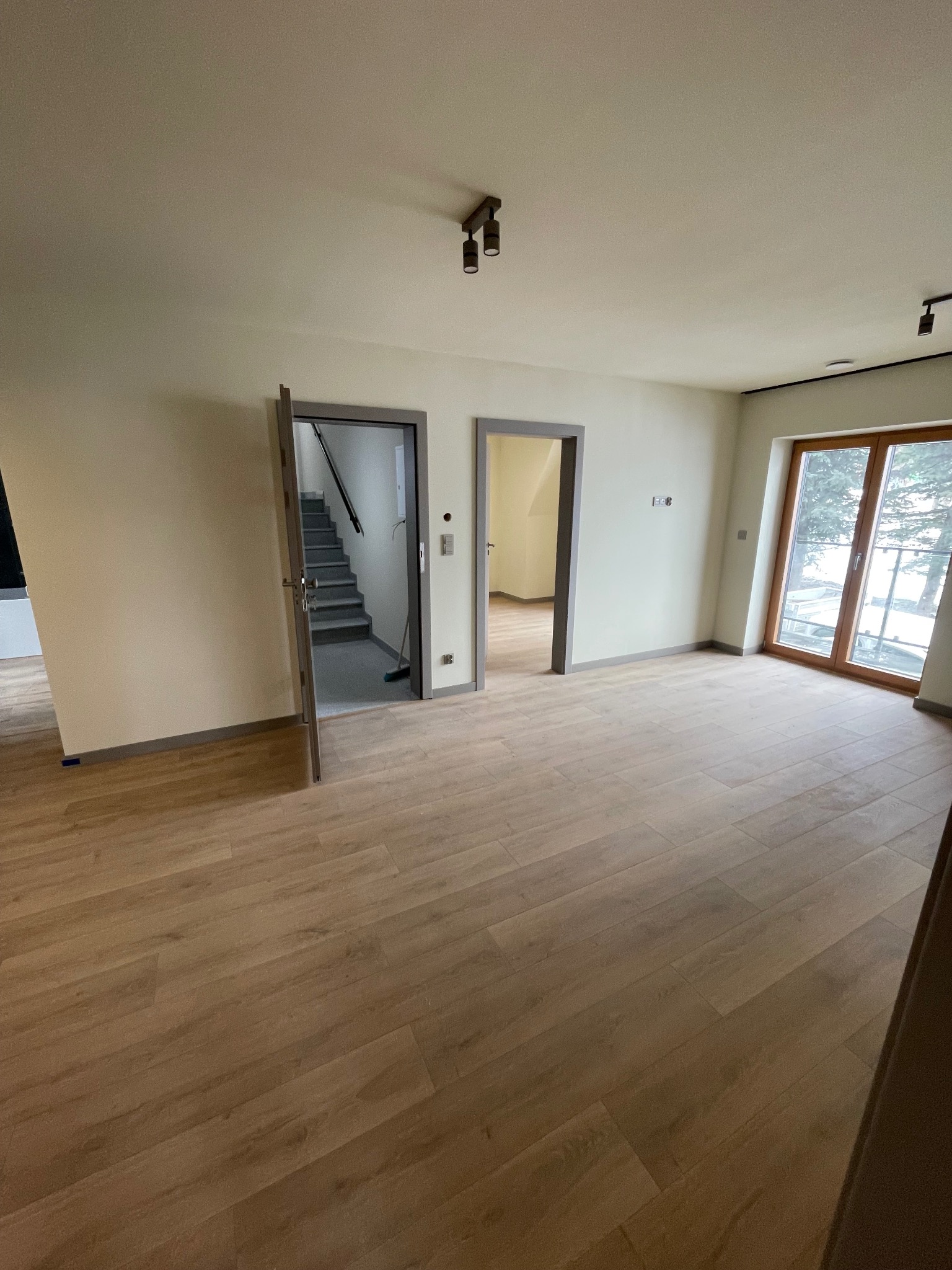 Jasne, przestronne wnętrze z drewnianą podłogą, widok na schody i okno z widokiem na zewnątrz. Minimalistyczny design, neutralna kolorystyka ścian, nowoczesne oświetlenie.