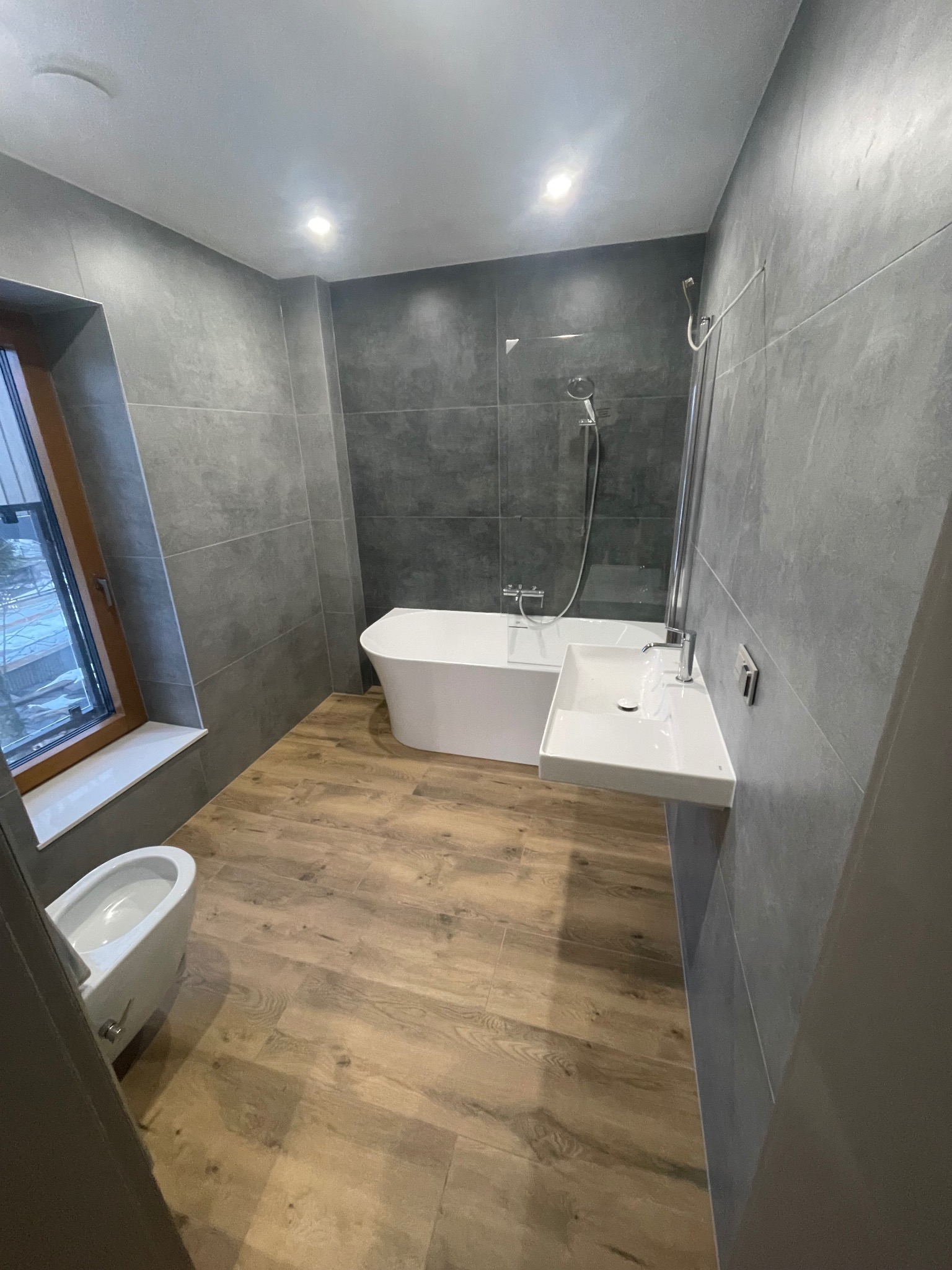 Nowoczesna łazienka z wolnostojącą wanną, wiszącą umywalką i bidetem. Szare płytki na ścianach kontrastują z drewnopodobną podłogą. Okno z widokiem na zewnątrz.