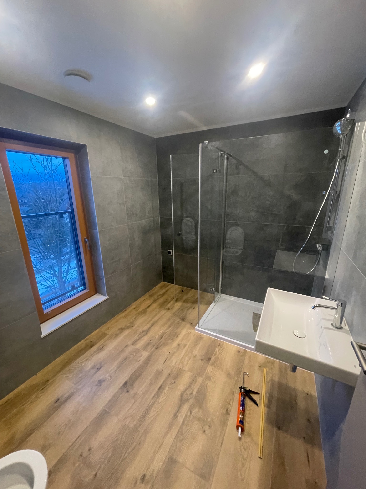 Nowoczesna łazienka z kabiną prysznicową, umywalką i drewnopodobną podłogą. Widoczne narzędzia do silikonowania. Za oknem zimowy krajobraz.