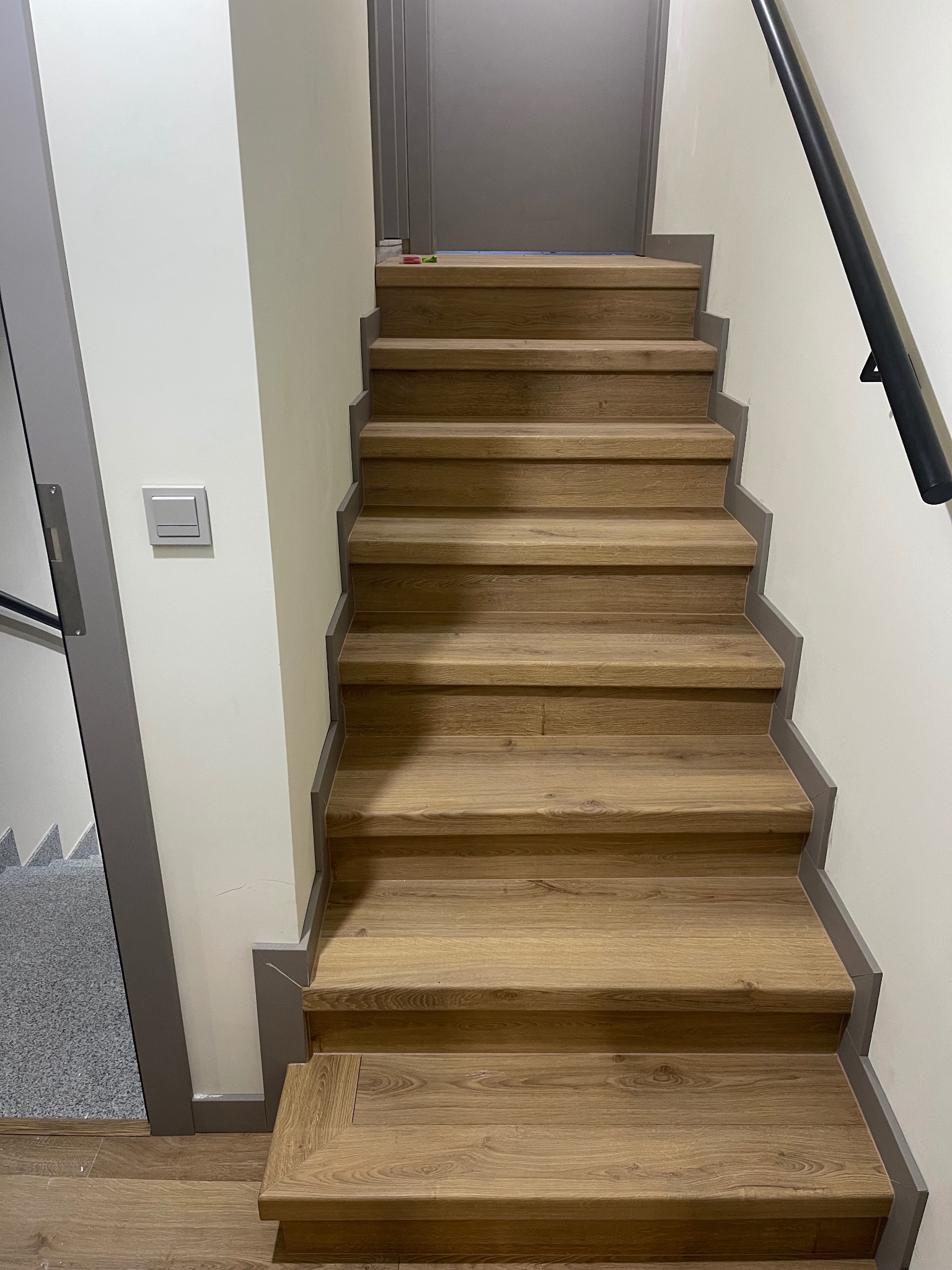 Drewniane schody z szarą listwą przypodłogową i poręczą, widok z dołu. Nowoczesne wykończenie wnętrza, jasne ściany i szare drzwi w tle.