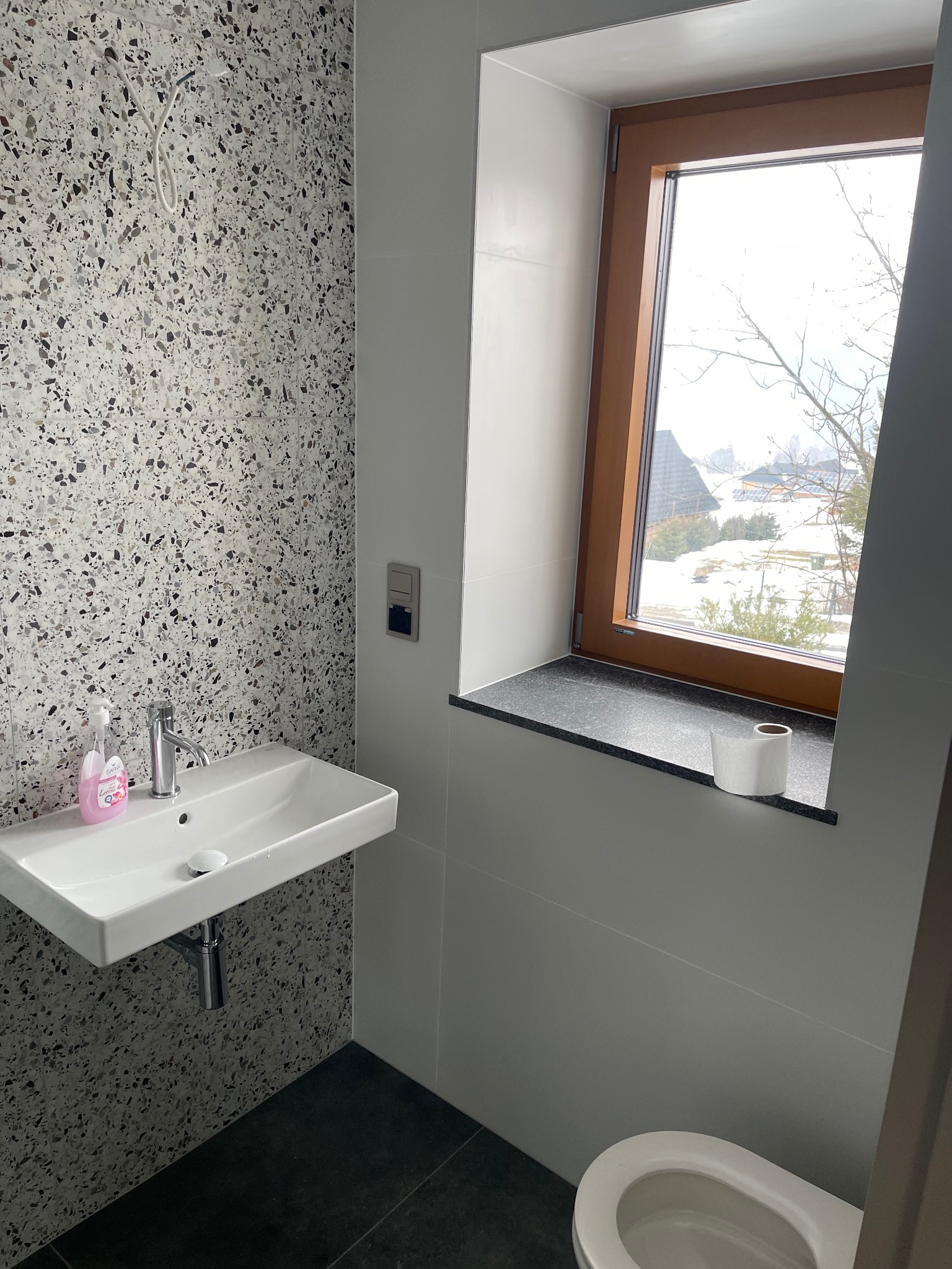 Nowoczesna łazienka z umywalką na tle ściany w terazzo, drewniane okno z widokiem na zimowy krajobraz. Minimalistyczny design, czarne płytki podłogowe.