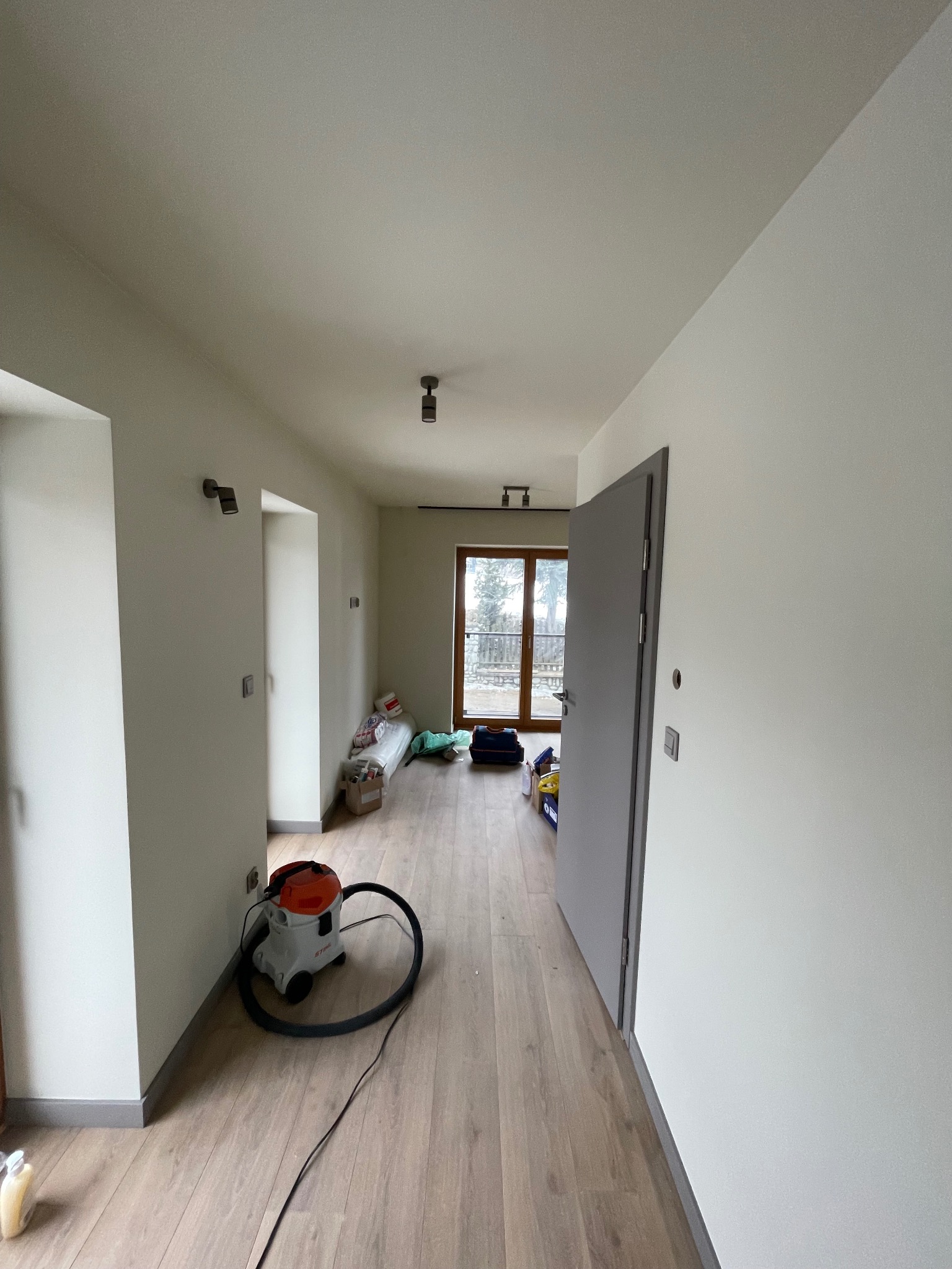 Jasne wnętrze po malowaniu ścian, widoczny odkurzacz przemysłowy, podłoga drewniana, szare drzwi i okno w tle. Minimalistyczne oświetlenie sufitowe.
