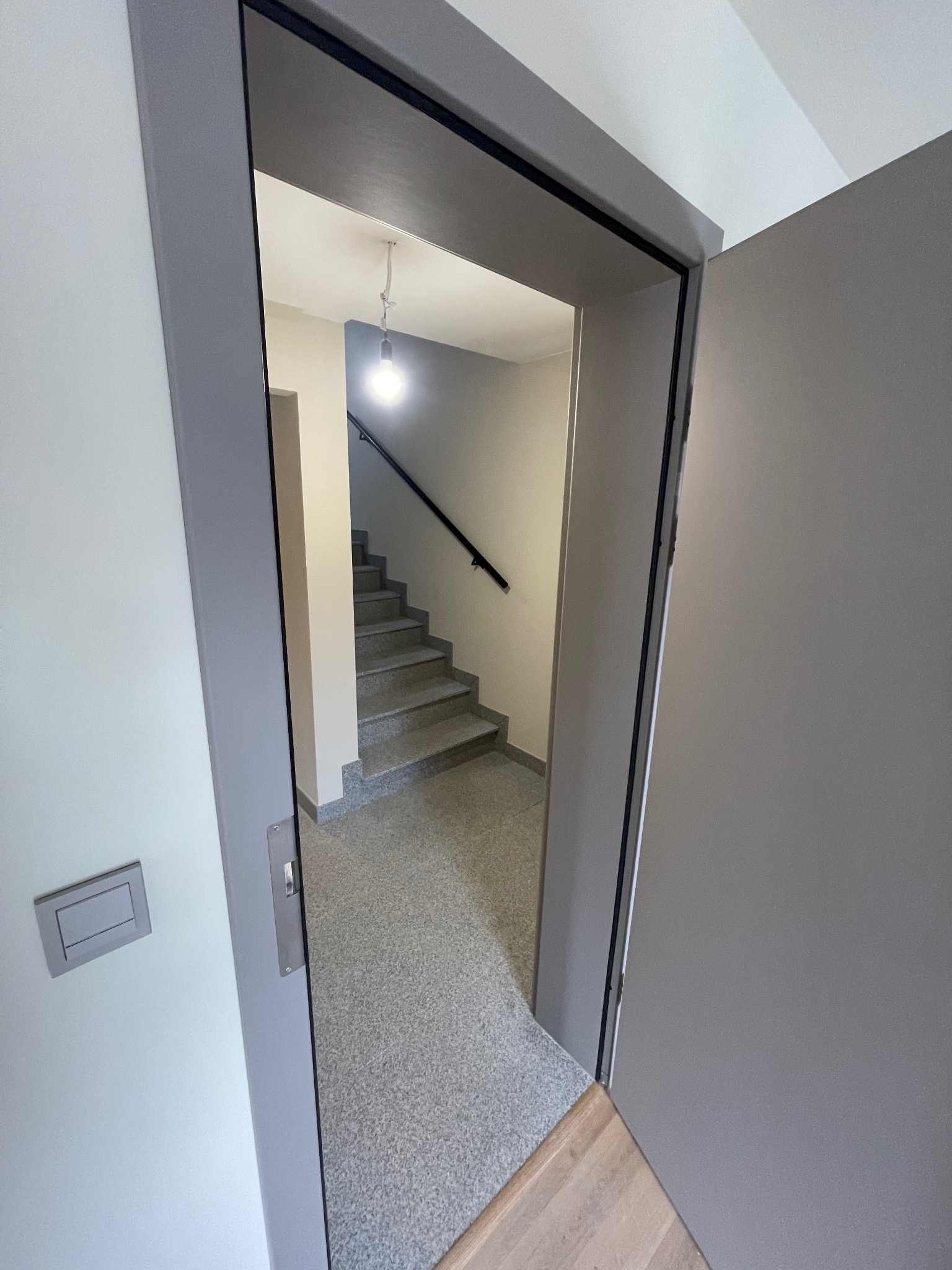 Otwarta szara, przesuwna ściana działowa ukazuje klatkę schodową z szarymi schodami i czarną poręczą. Widoczna lampa sufitowa i fragment drewnianej podłogi.